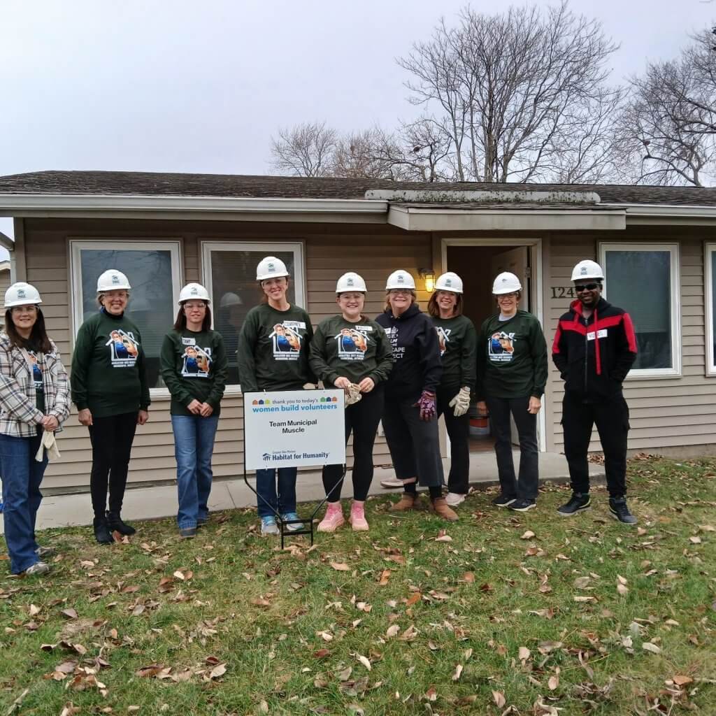 A group of women smiling in front of a house with construction hats on.