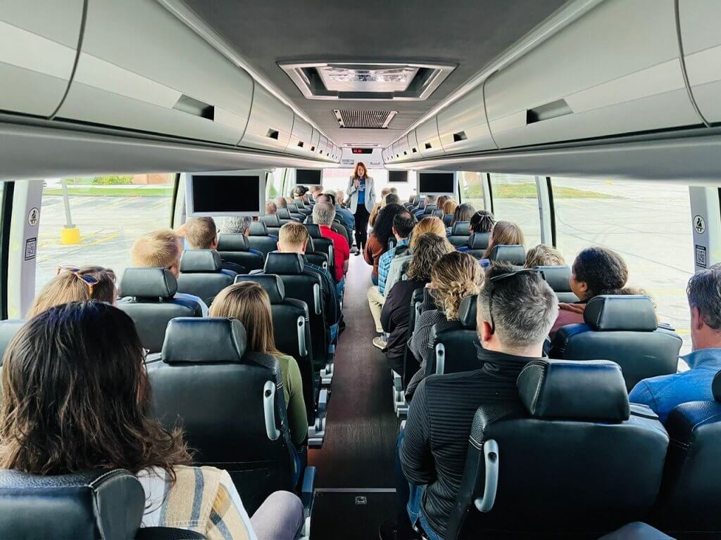A group on a bus with a woman walking down the center holding a microphone.