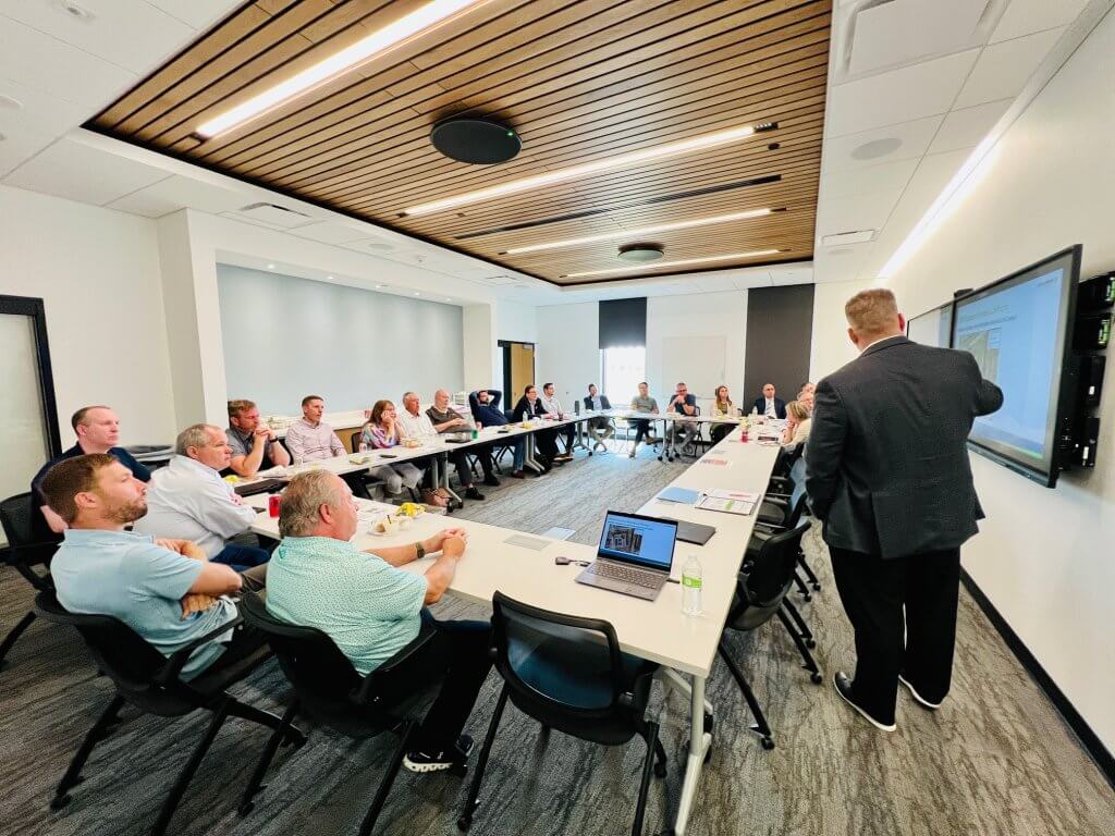 A man presenting a powerpoint on a screen with a group of business professionals sitting around a table listening and watching.