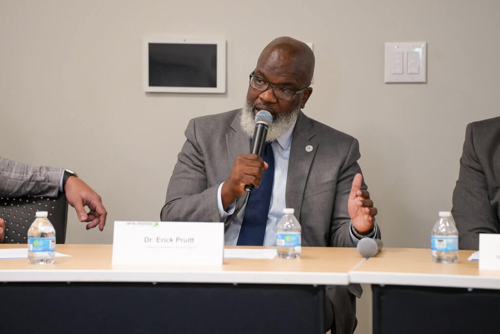 A man holding a microphone and speaking. He has a nametag in front of him that reads "Dr. Erick Pruitt"