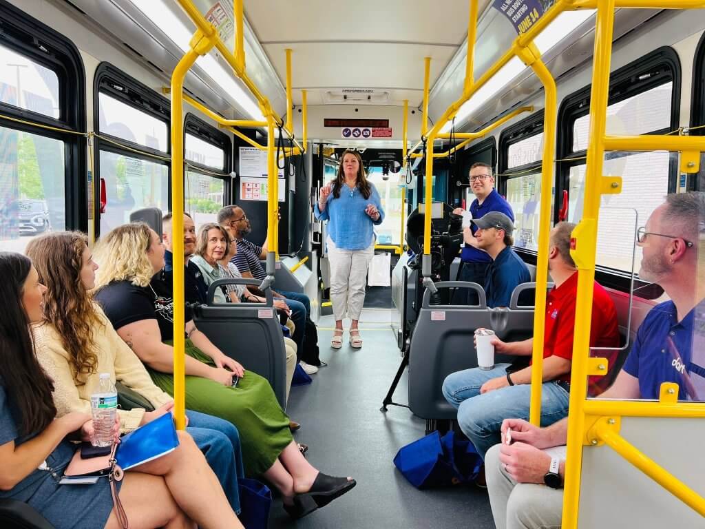 People on a bus smiling and listening to a woman talk at the front.