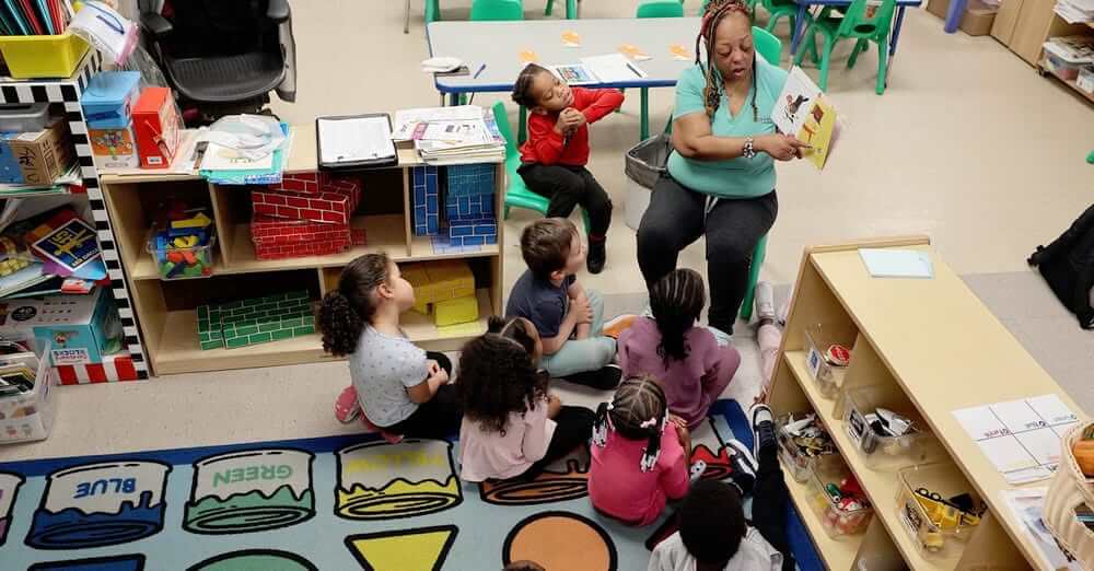 Children watching and listening while a woman reads them a picture book.