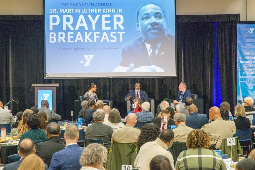 A group of people watching three individuals on stage with a graphic for Dr Martin Luther King Jr. Prayer Breakfast